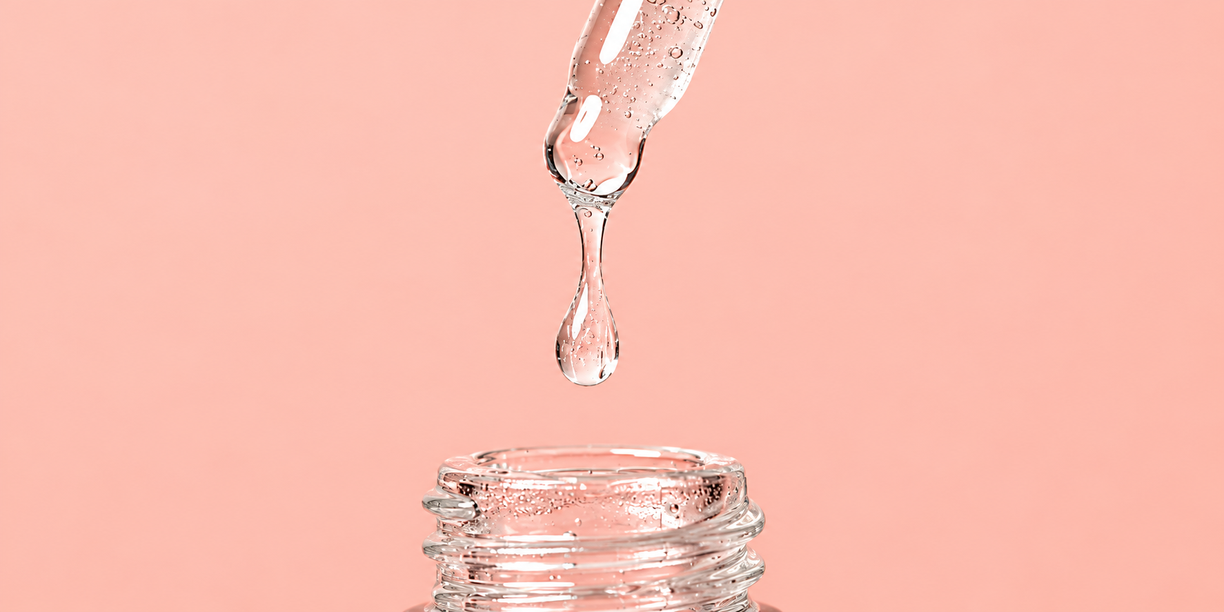 Clear serum dripping from a glass dropper into an open bottle on a peach background